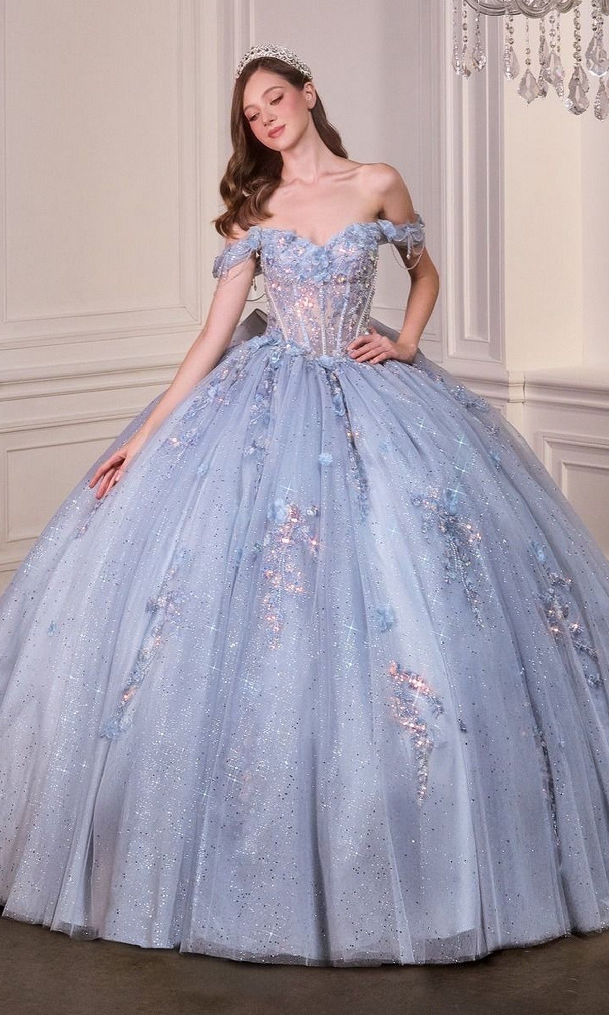 A woman wearing the Quinceanera Dress 15728 By Ladivine, a glitter tulle light blue off-the-shoulder gown, and a tiara stands in an elegant room with white walls.